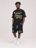 A man modeling a black athletic t-shirt and matching shorts set with gold and white graphics, paired with white socks and sneakers, against a light gray studio background.
