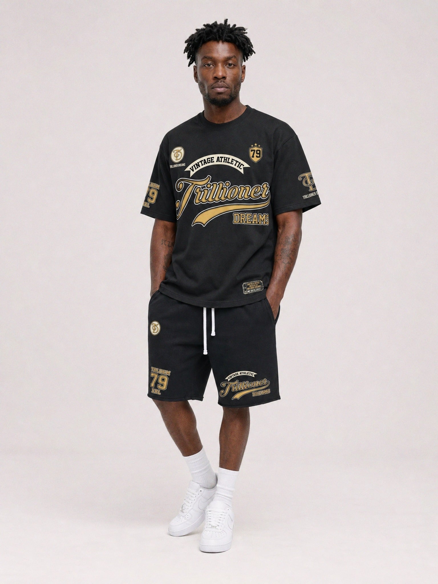 A man modeling a black athletic t-shirt and matching shorts set with gold and white graphics, paired with white socks and sneakers, against a light gray studio background.