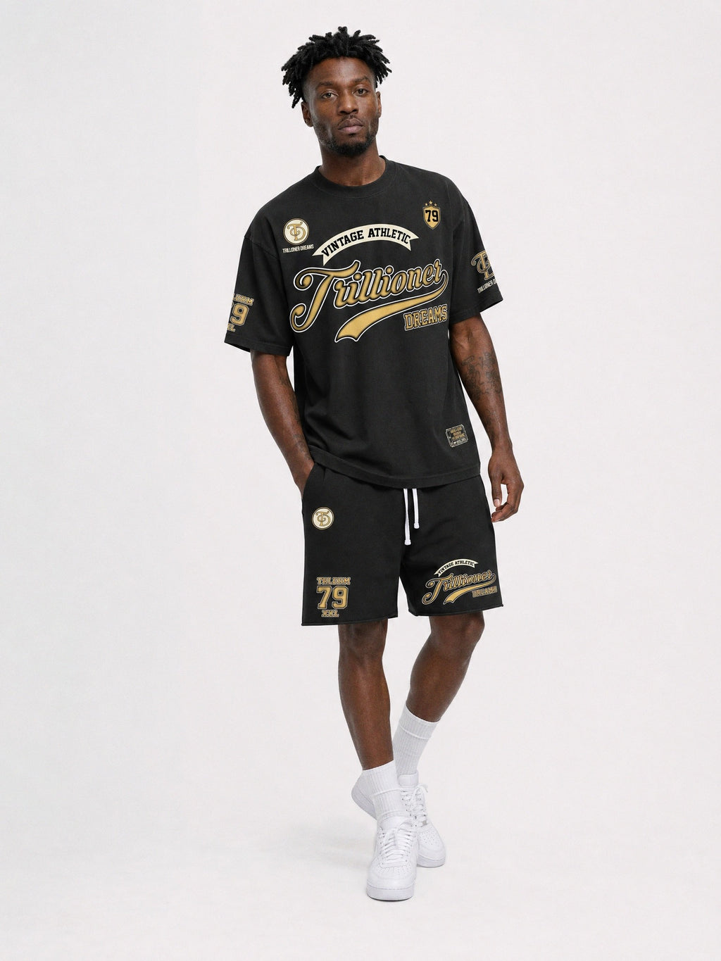 A man modeling a black athletic t-shirt and matching shorts set with gold and white graphics, paired with white socks and sneakers, against a plain light gray studio background.