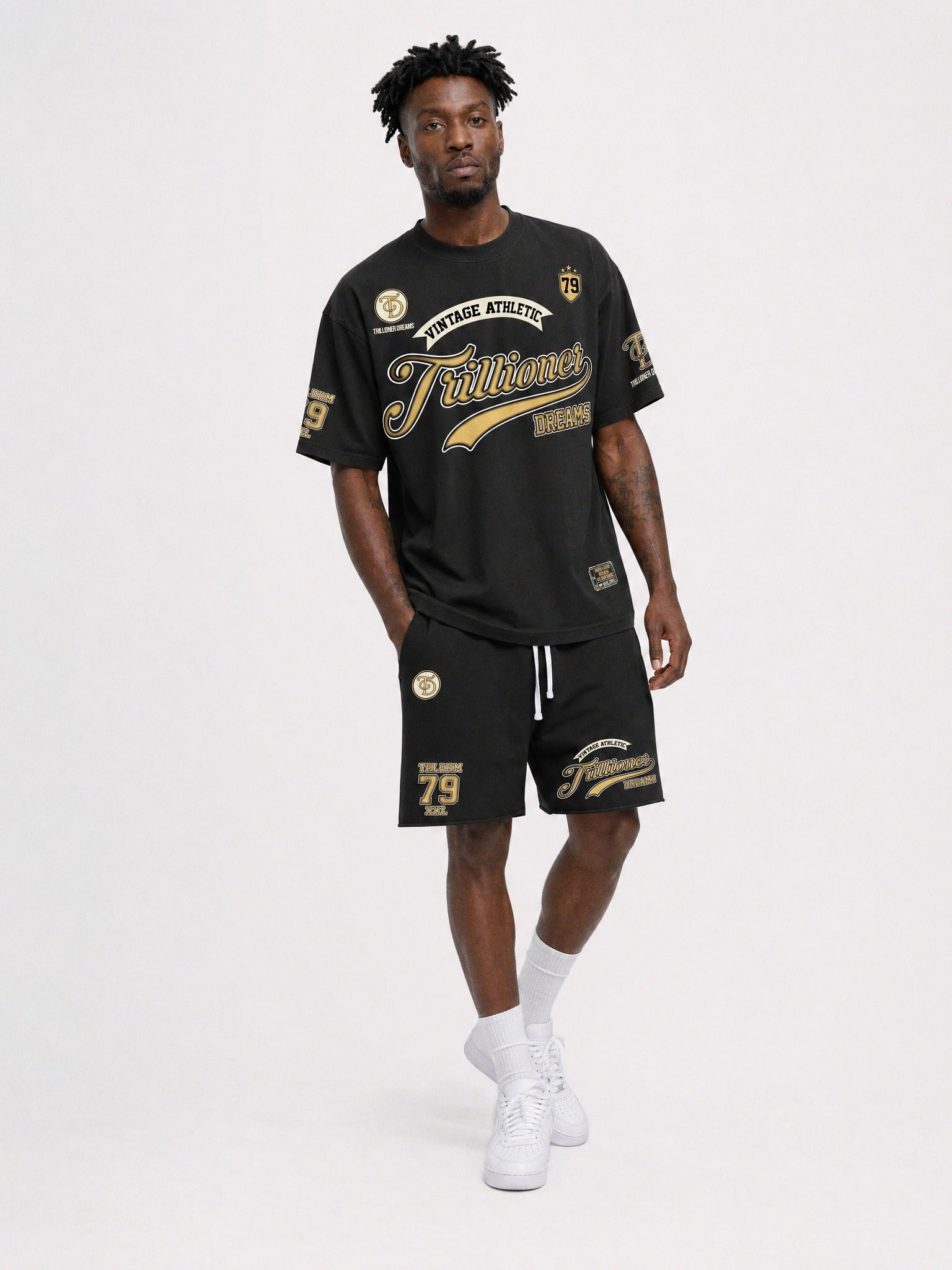 A man modeling a black athletic t-shirt and matching shorts set with gold and white graphics, paired with white socks and sneakers, against a plain light gray studio background.