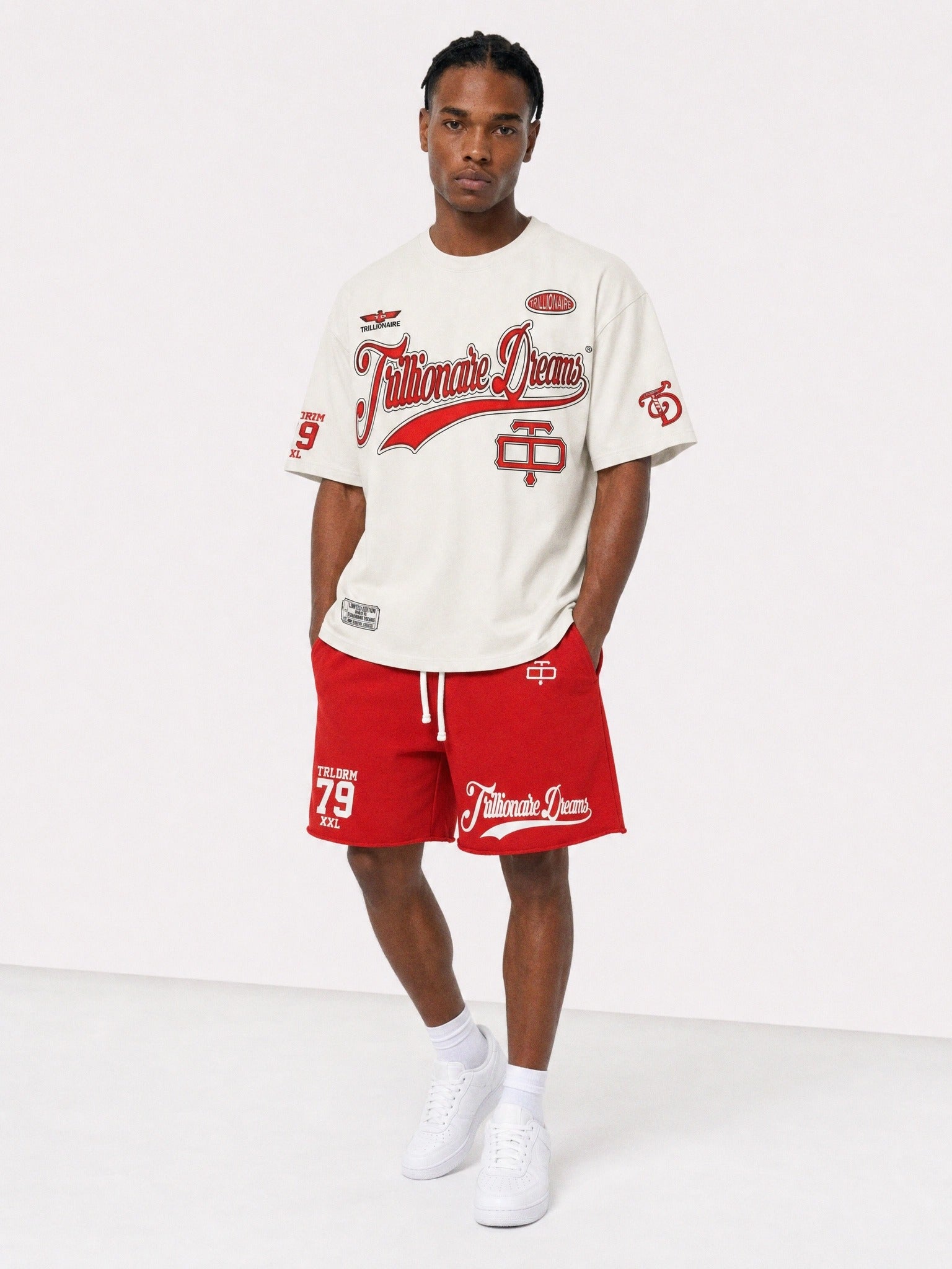 A male model stands against a plain light gray studio background wearing a cream graphic T-shirt and bright red athletic shorts with white graphics and text, plus white sneakers.