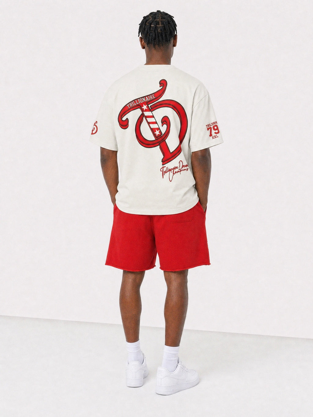 A man viewed from behind wearing a cream-colored t-shirt with a large red graphic on the back, paired with red shorts, white socks, and white sneakers, set against a plain white studio background.