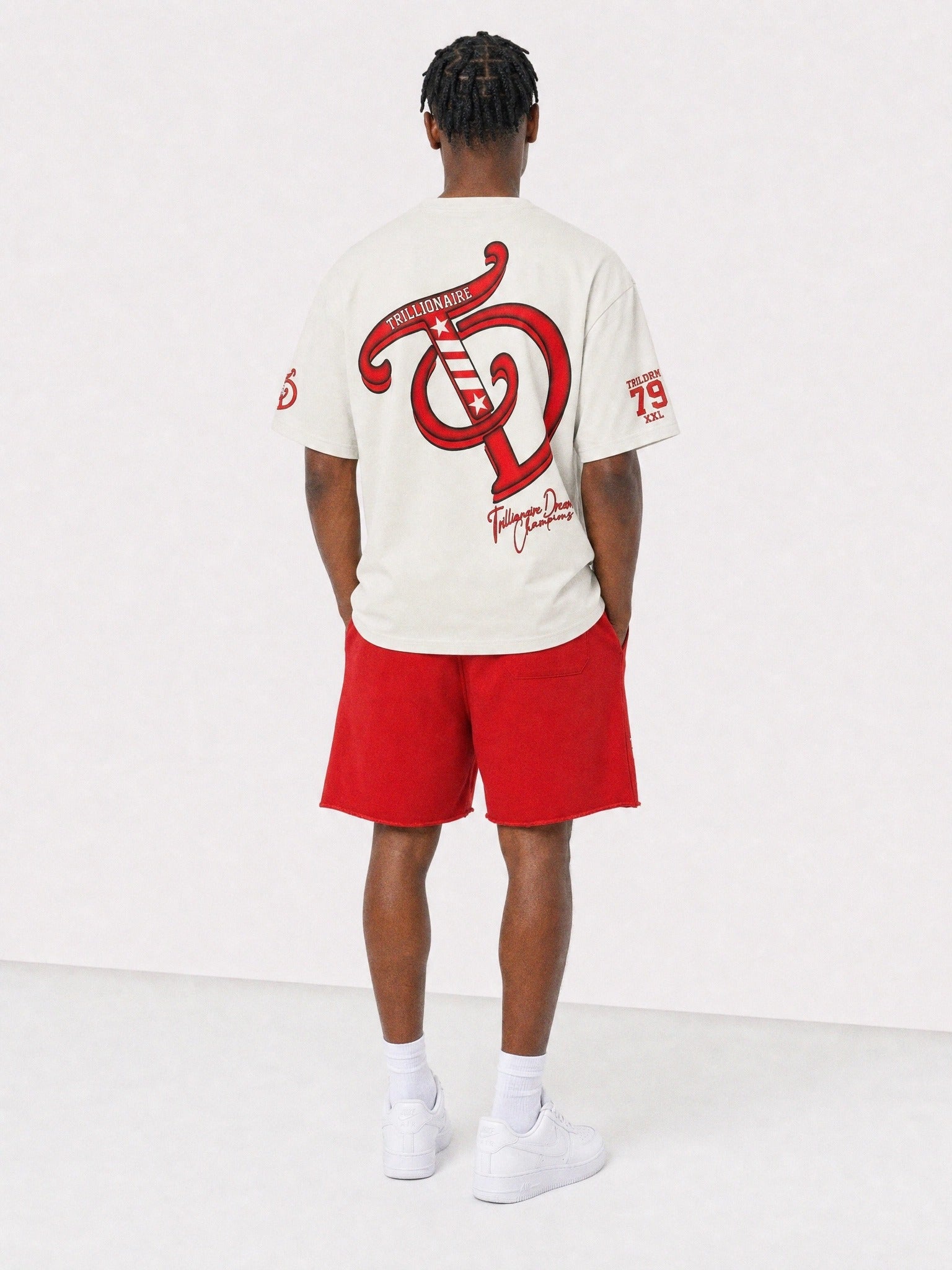 A man viewed from behind wearing a cream-colored t-shirt with a large red graphic on the back, paired with red shorts, white socks, and white sneakers, set against a plain white studio background.