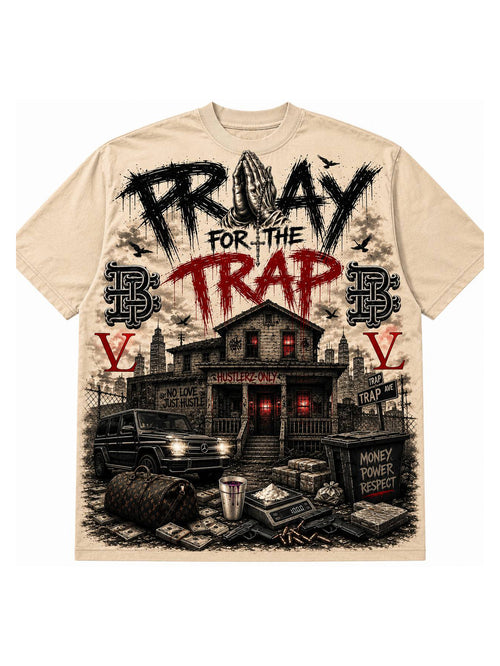 A beige short-sleeve t-shirt with a large graphic print on the front. The design features the text 'PRAY FOR THE TRAP' in black and red distressed lettering above a detailed black and red illustration of a trap house, a vehicle, cash, and stacks of money.